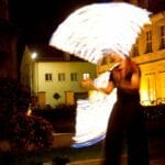 Feuerkünstler begeistert mit einer Hochzeitsfeuershow, bei der er mithilfe eines Feuerseils helle, wirbelnde Lichtspuren vor beleuchteten Gebäuden erzeugt.