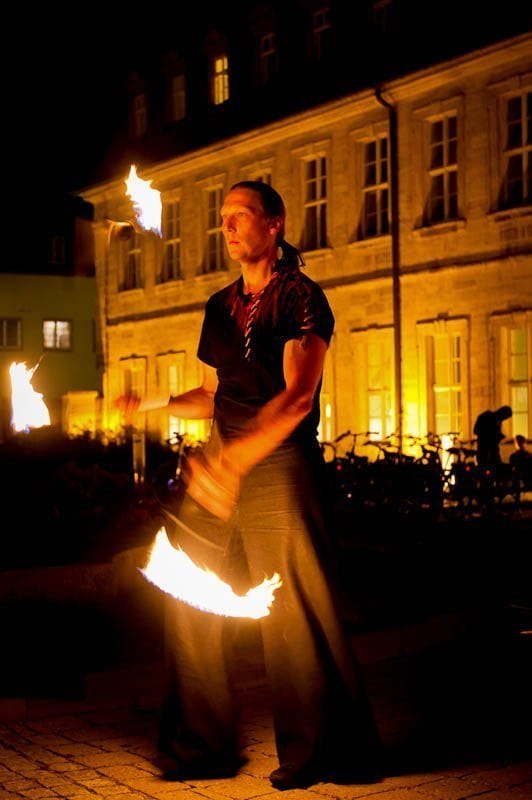 Detlef Vogt führt eine Feuershow auf, indem sie nachts vor einem wunderschön beleuchteten Gebäude mit brennenden Fackeln jongliert und so für eine unvergessliche Hochzeitsfeier sorgt.